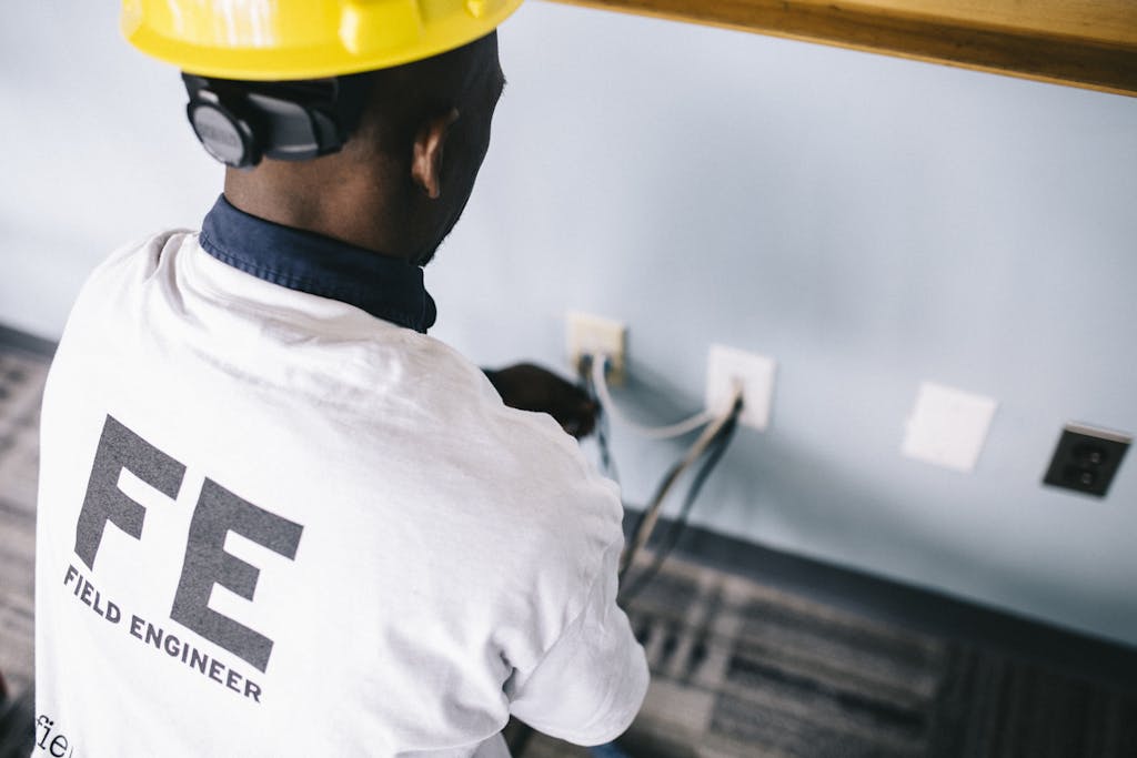 From above back view unrecognizable black field engineer wearing white shirt and protective hardhat sitting on floor and checking voltage in wall plugs